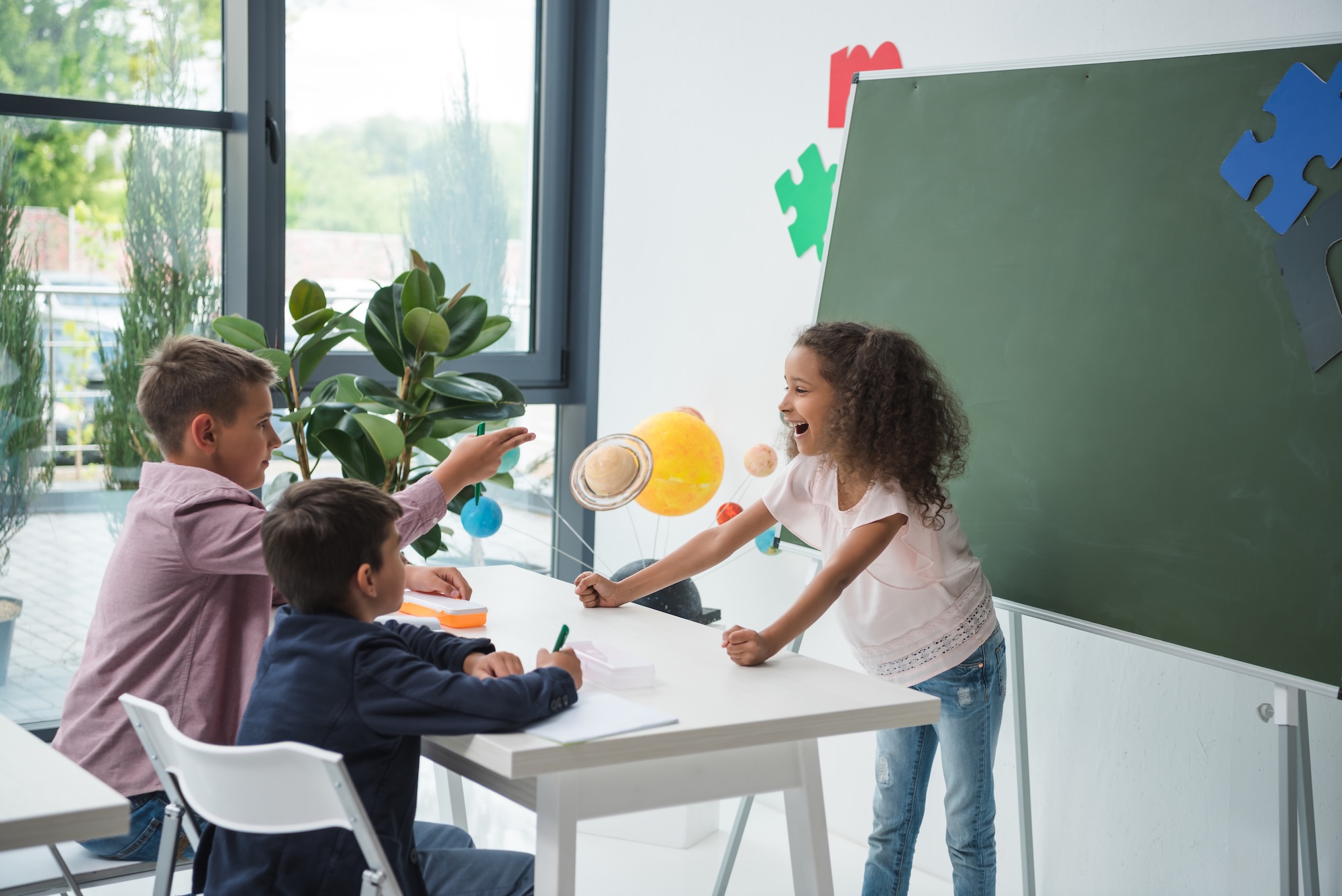 Adorable multiethnic schoolchildren talking and smiling at desk in classroom Smiling children in a classroom engage in a lively discussion near a chalkboard and solar system model.