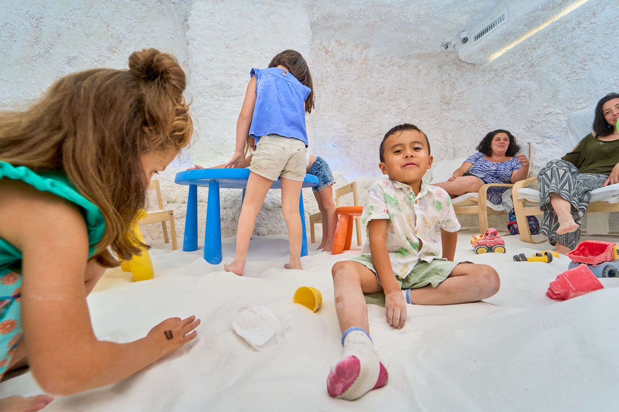 Children playing with toys in an indoor sand-covered room Children play in an indoor sensory room with soft sand and toys while adults observe nearby.