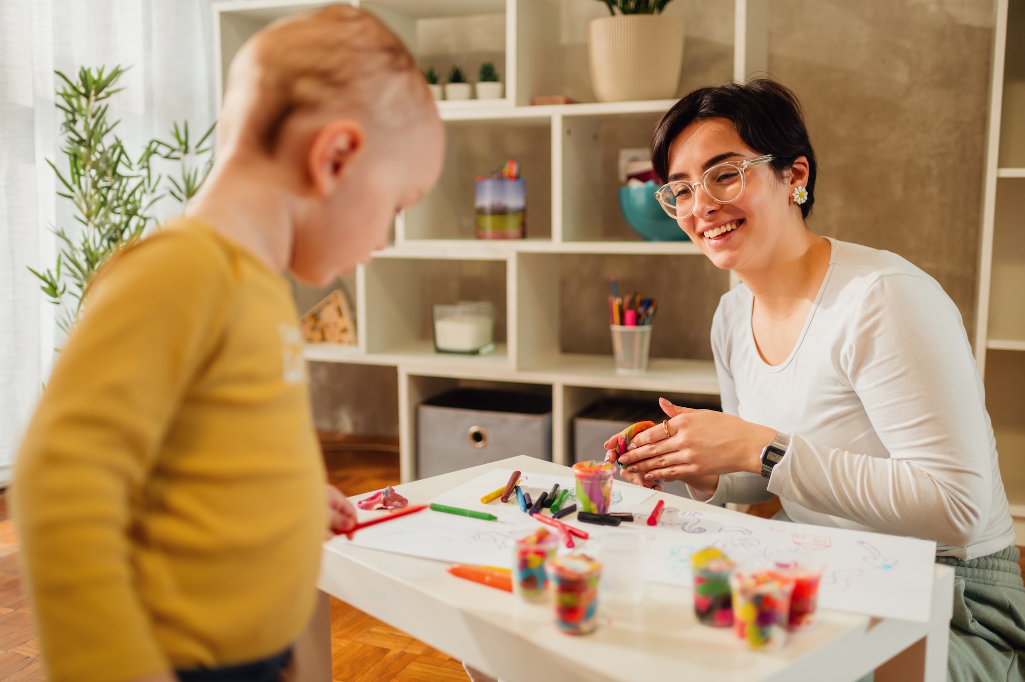 Mother and son drawing with crayons at home and creating art A smiling therapist sits at a small table doing art activities with a young child during an ABA therapy session.